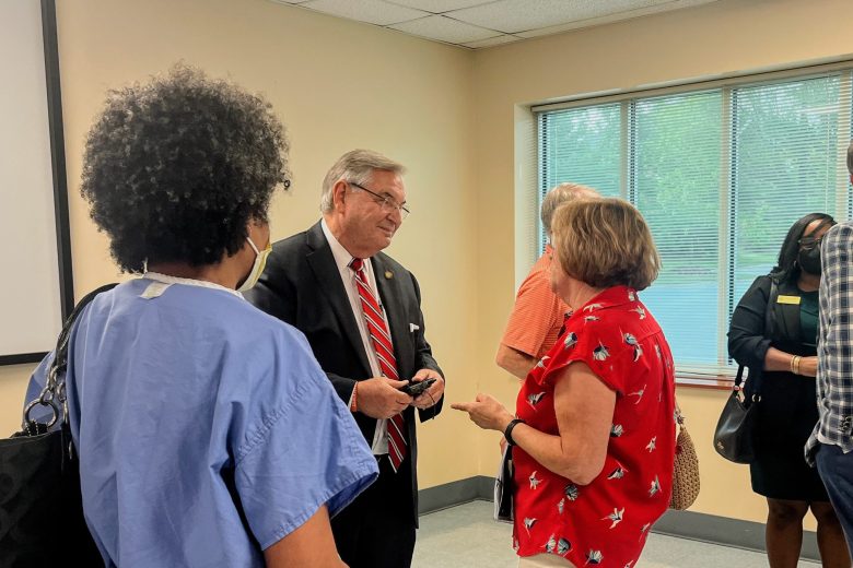 older white man in suit speaks to an older white woman while a Black woman in medical scrubs looks on
